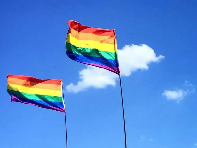Zwei LQBTQ-Flaggen flattern im Wind. Im Hintergrund ist blauer Himmel mit Sonnenschein und zwei kleine Wolken zu sehen.