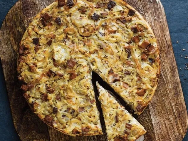 von Lena Marie Radu - Ein runder Zwiebelkuchen auf einem Holzbrett mit einem herausgeschnittenen Stück auf einer Tortenschaufel.