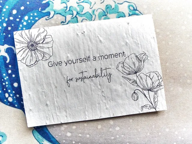 Eine cremefarbene Postkarte mit den Worten "Give yourself a moment for sustainability" liegt auf einem Kissen mit Wellenmotiv.