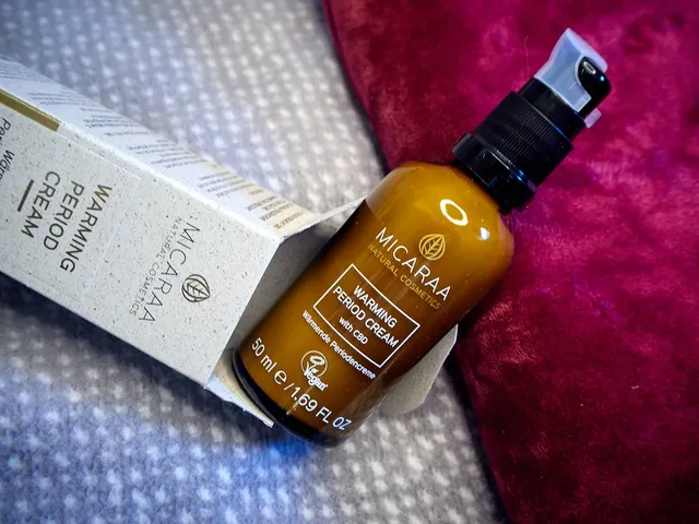 Eine Wärmflasche mit einer lila Samthülle liegt mit einem Teebeutel und einer beigen Verpackung für eine wärmende Periodencreme mit goldener Beschriftung auf einer grauen Decke mit weißen Punkten