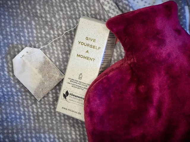 Eine Wärmflasche mit einer lila Samthülle liegt mit einem Teebeutel und einer beigen Verpackung für eine wärmende Periodencreme mit goldener Beschriftung auf einer grauen Decke mit weißen Punkten
