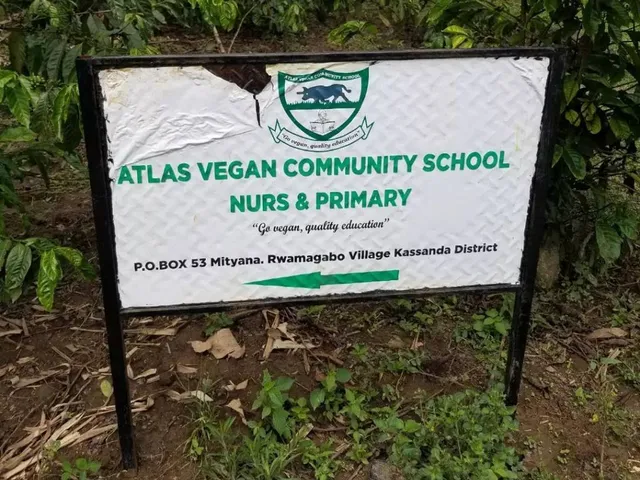 Ein schwarzes Metallschild steht auf einer Wiese mit Pflanzen im Hintergrund. Die grüne Aufschrift auf weißem Grund lautet: "Atlas Vegan Community School Nurs & Primary". Darüber ist das Logo der Schule zu sehen: ein Emblem mit einem vierbeinigen Tier und einer lesenden Person