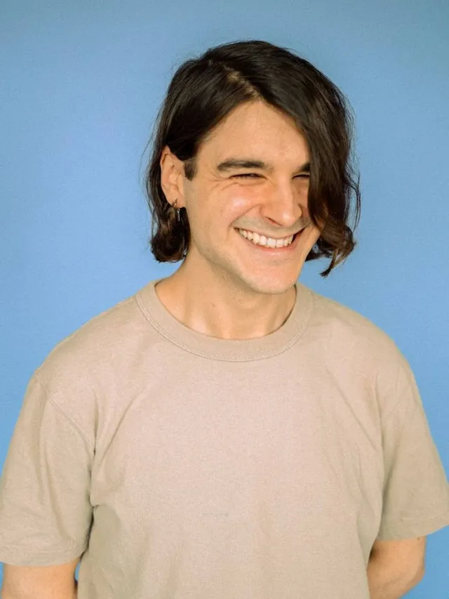 Simon steht lachend vor einem blauen Hintergrund. Er trägt ein hellgraues -Shirt und hat braune, halblange Haare.