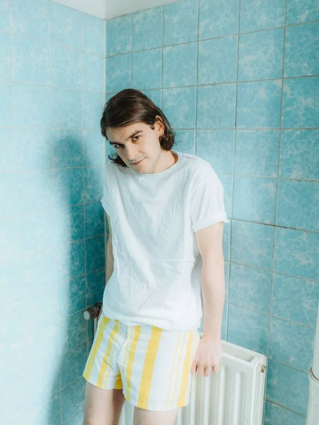 Simon Dübell steht an einen weißen Heizkörper gelehnt vor einer türkisfarbenen Fliesenwand und schaut nachdenklich in die Kamera. Er trägt gelbe Boxershorts und ein weißes T-Shirt und hat braune, halblange Haare.